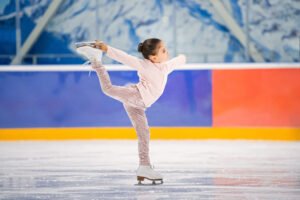 little girl figure skater in a light pink tracksuit with a smile skates on the ice on an indoor skating rink.